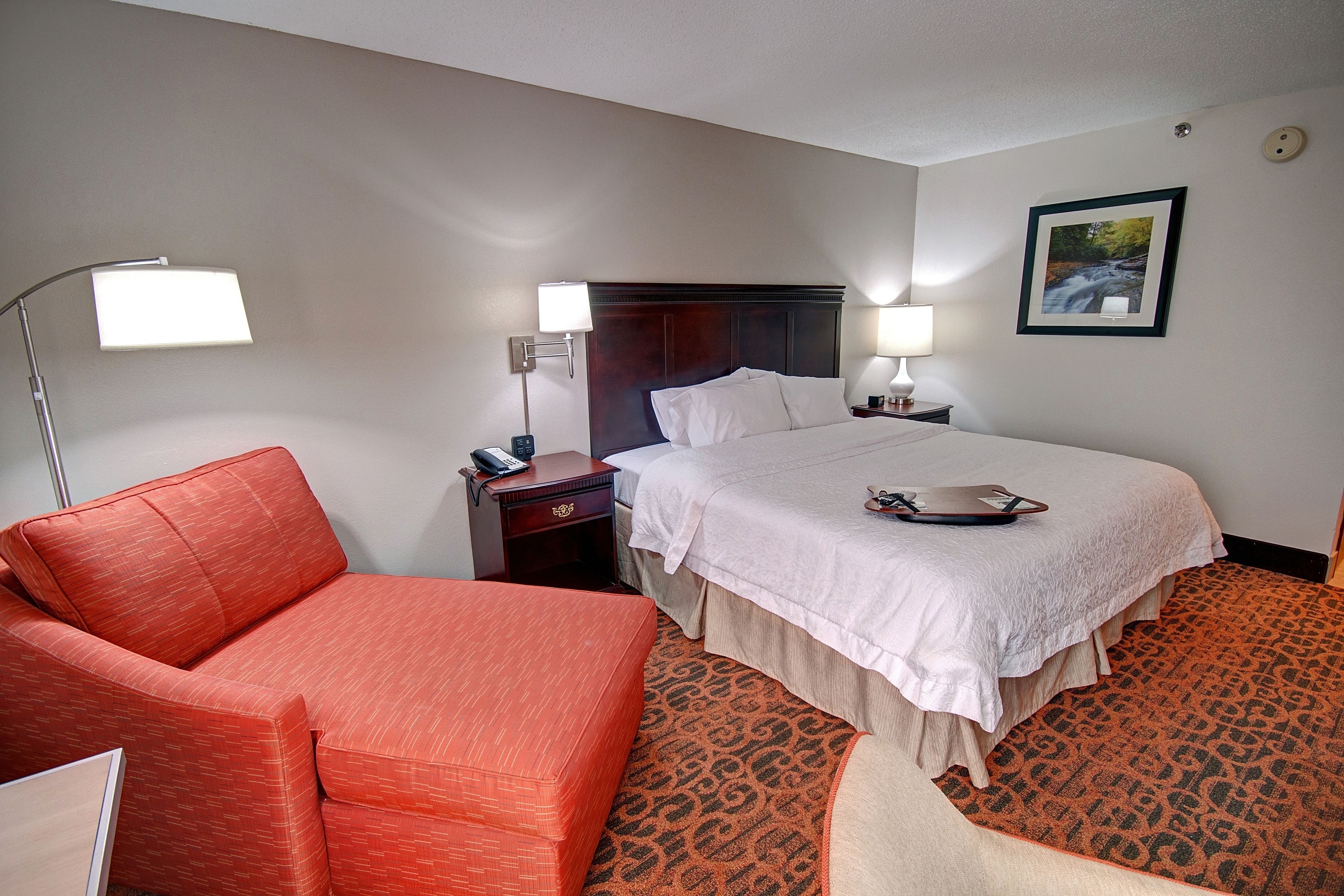 Habitación de hotel con cama, un sofá rojo, una mesita de noche, dos lámparas de noche y un cuadro en la pared.