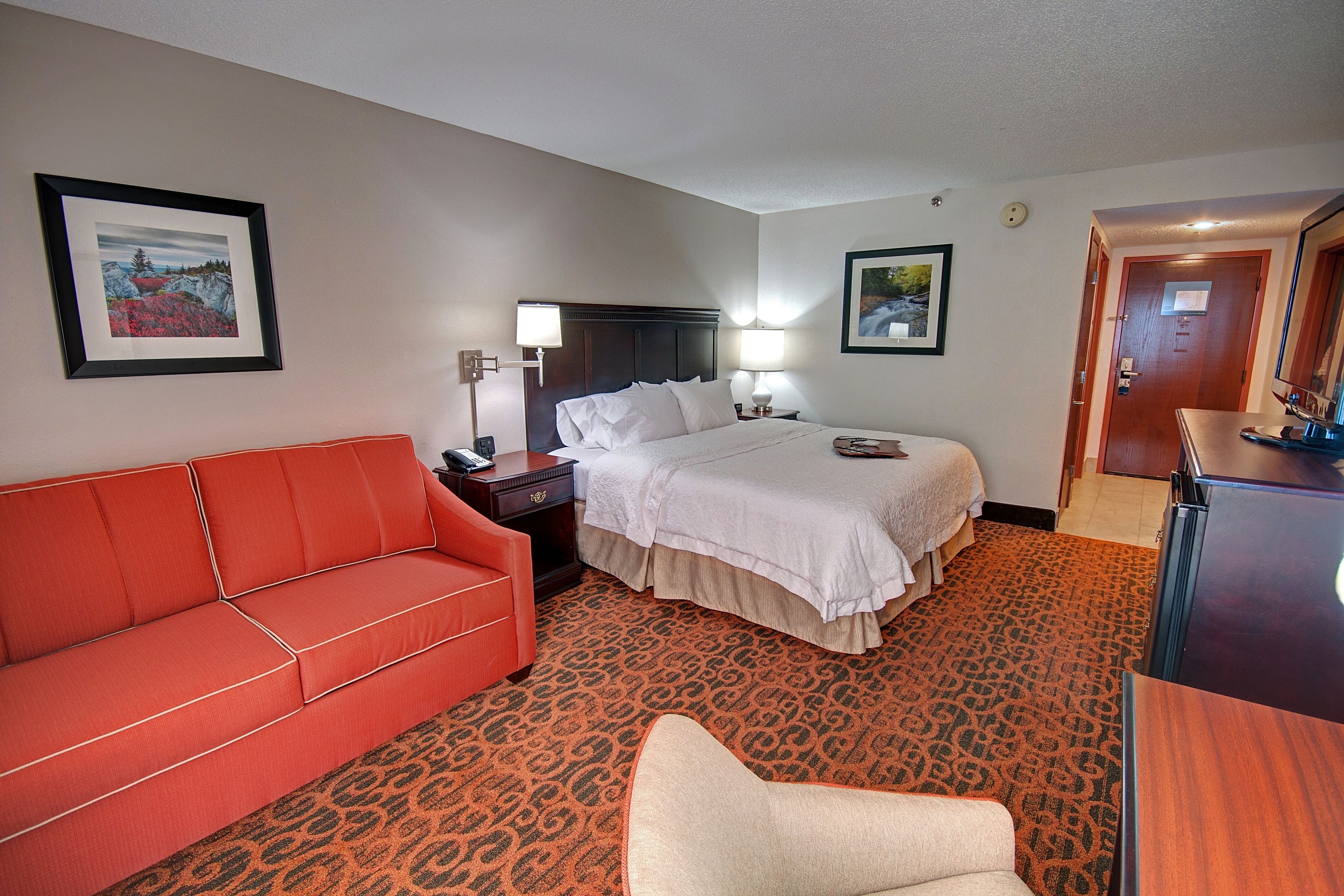 A hotel room with a bed, a red sofa, a small armchair, a nightstand, and a desk.