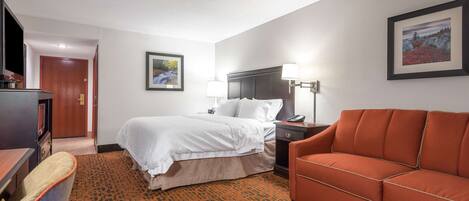 A hotel room with a bed, a sofa, a desk, a lamp, and a framed picture on the wall.