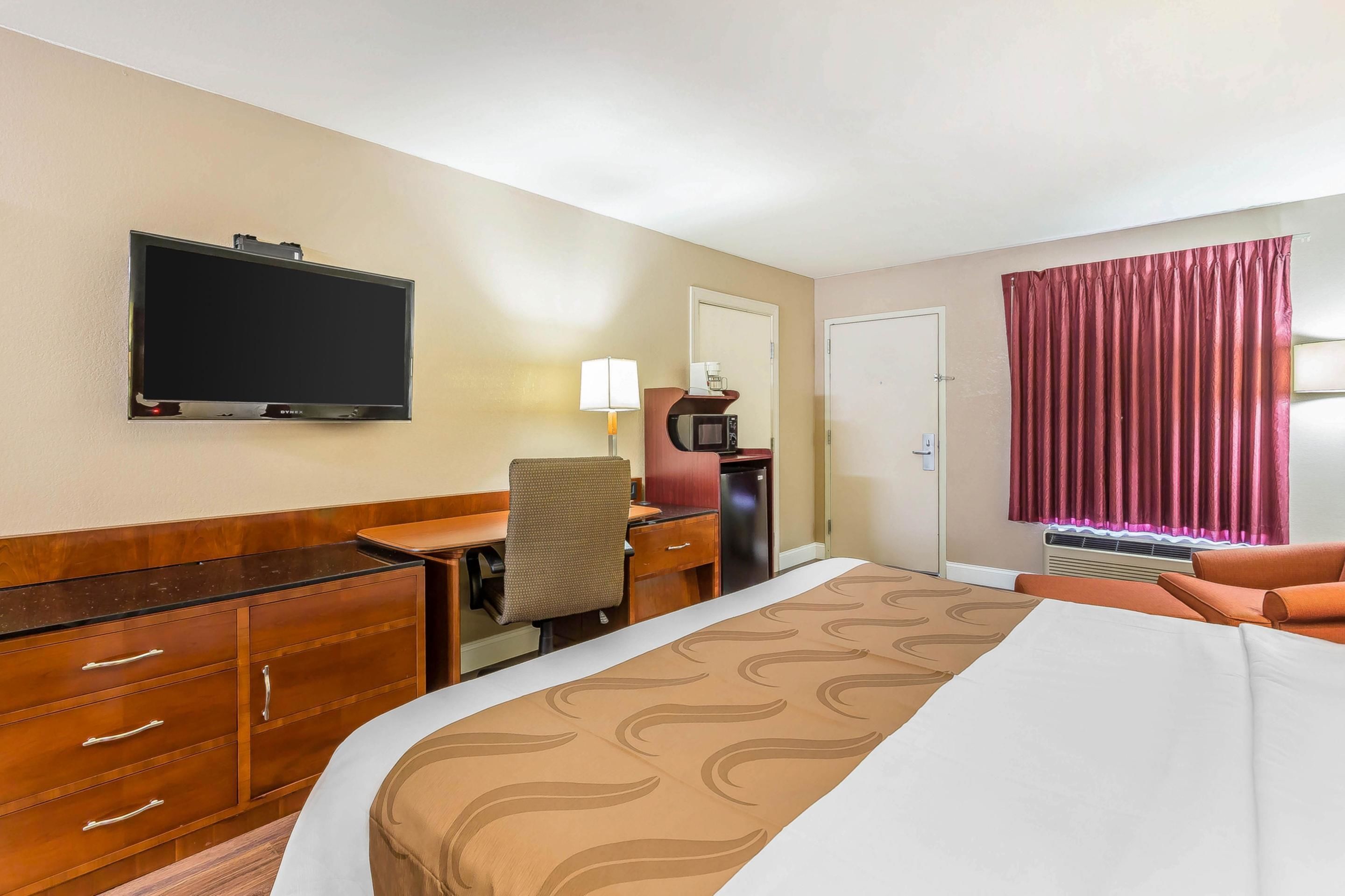 A hotel room with a bed, a desk with a chair, a television, a microwave, a lamp, and a window with curtains.