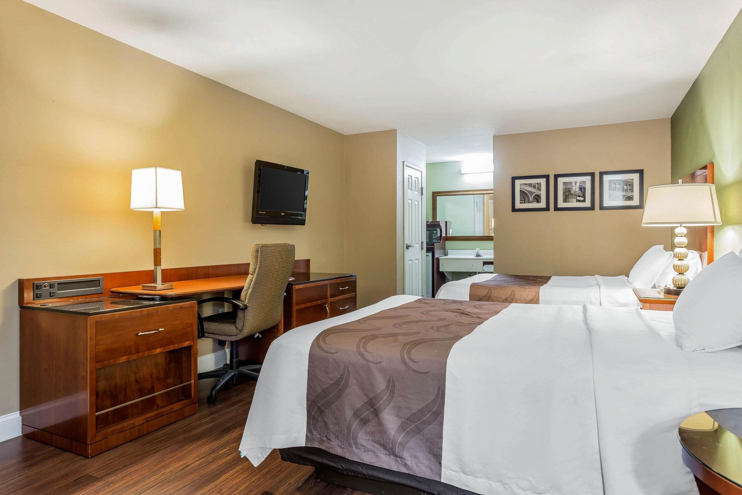 Une chambre d’hôtel avec un bureau en bois, deux lits, une chaise, une télévision et une porte donnant sur une salle de bain.