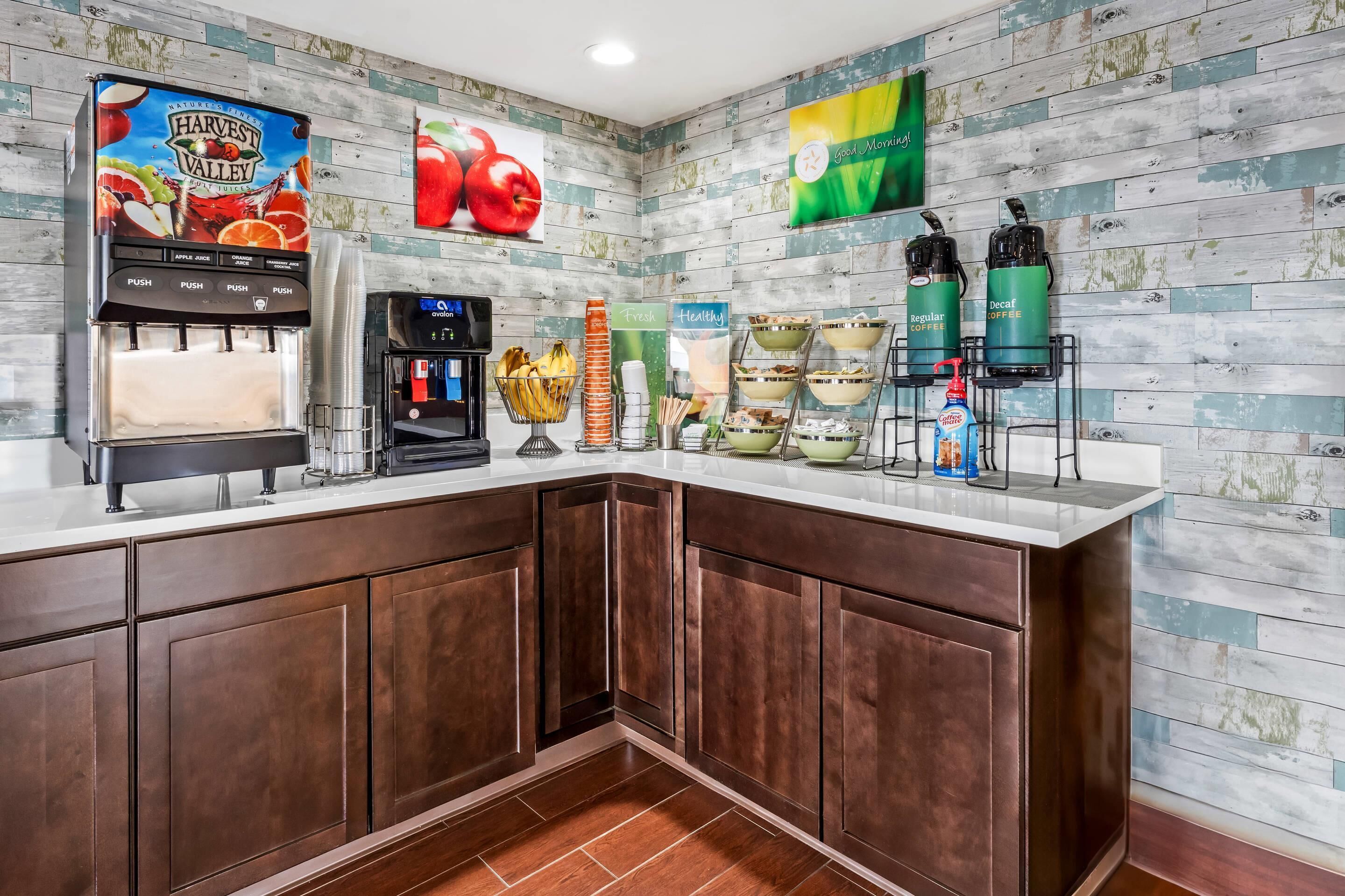 A self-service beverage station with a juice dispenser, coffee machines, and a fresh fruit display.