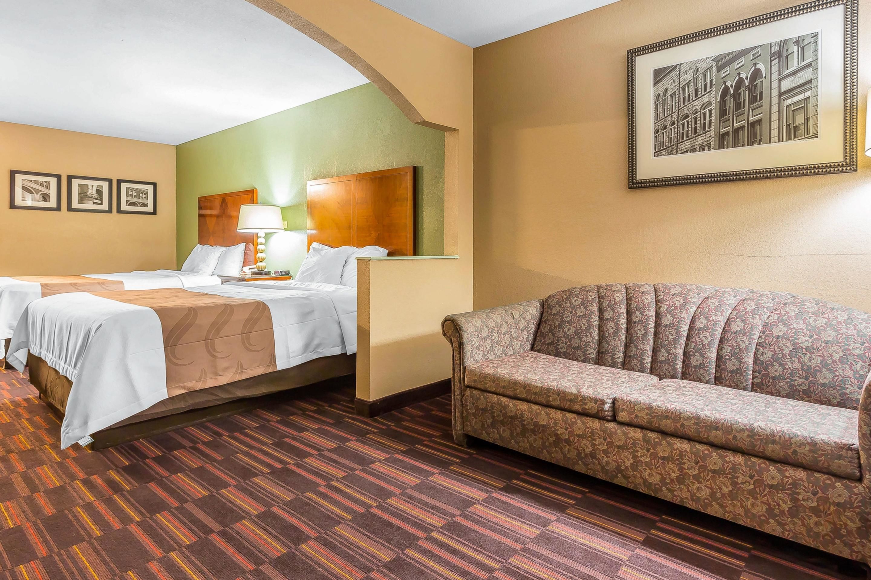 A hotel room with a bed, a sofa, a lamp, and framed pictures on the wall.