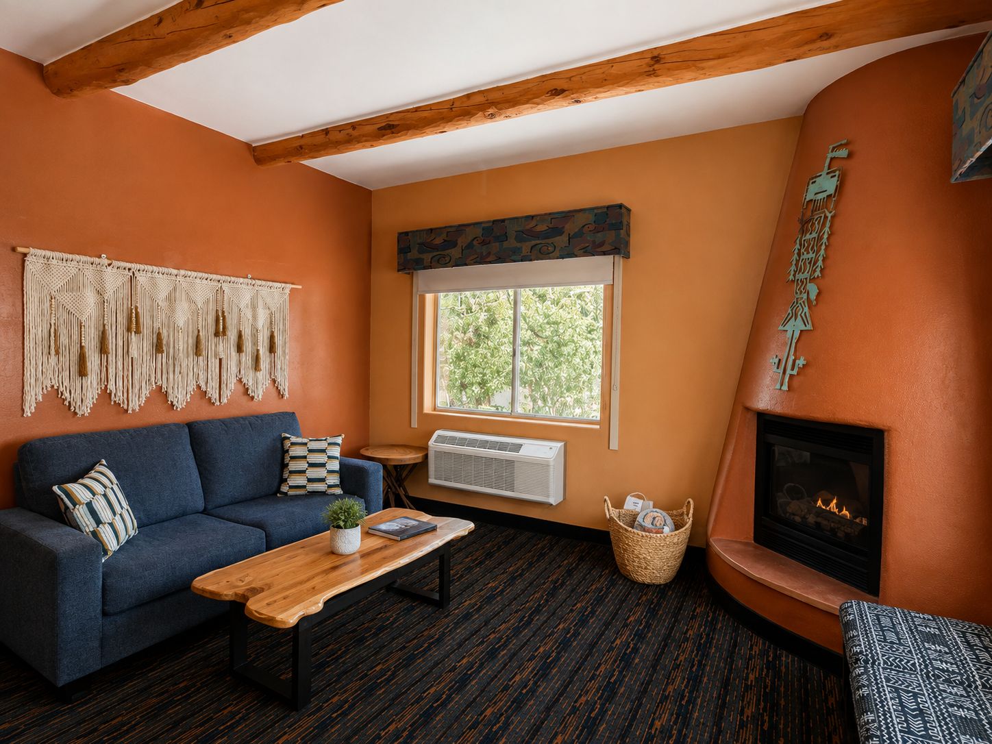 A cozy living room with a blue sofa, a wooden coffee table, a fireplace, and a large window with a view of trees.