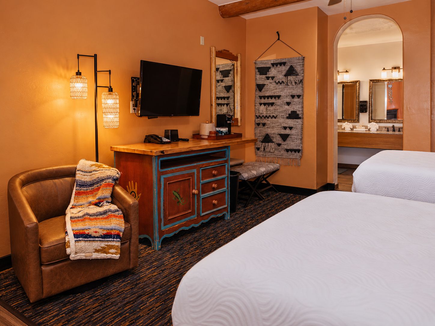 A hotel room with a bed, a desk with a flat-screen TV, a chair with a blanket, a hanging tapestry, and a bathroom mirror.