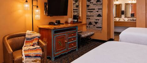 A hotel room with a bed, a desk with a flat-screen TV, a chair with a blanket, a hanging tapestry, and a bathroom mirror.