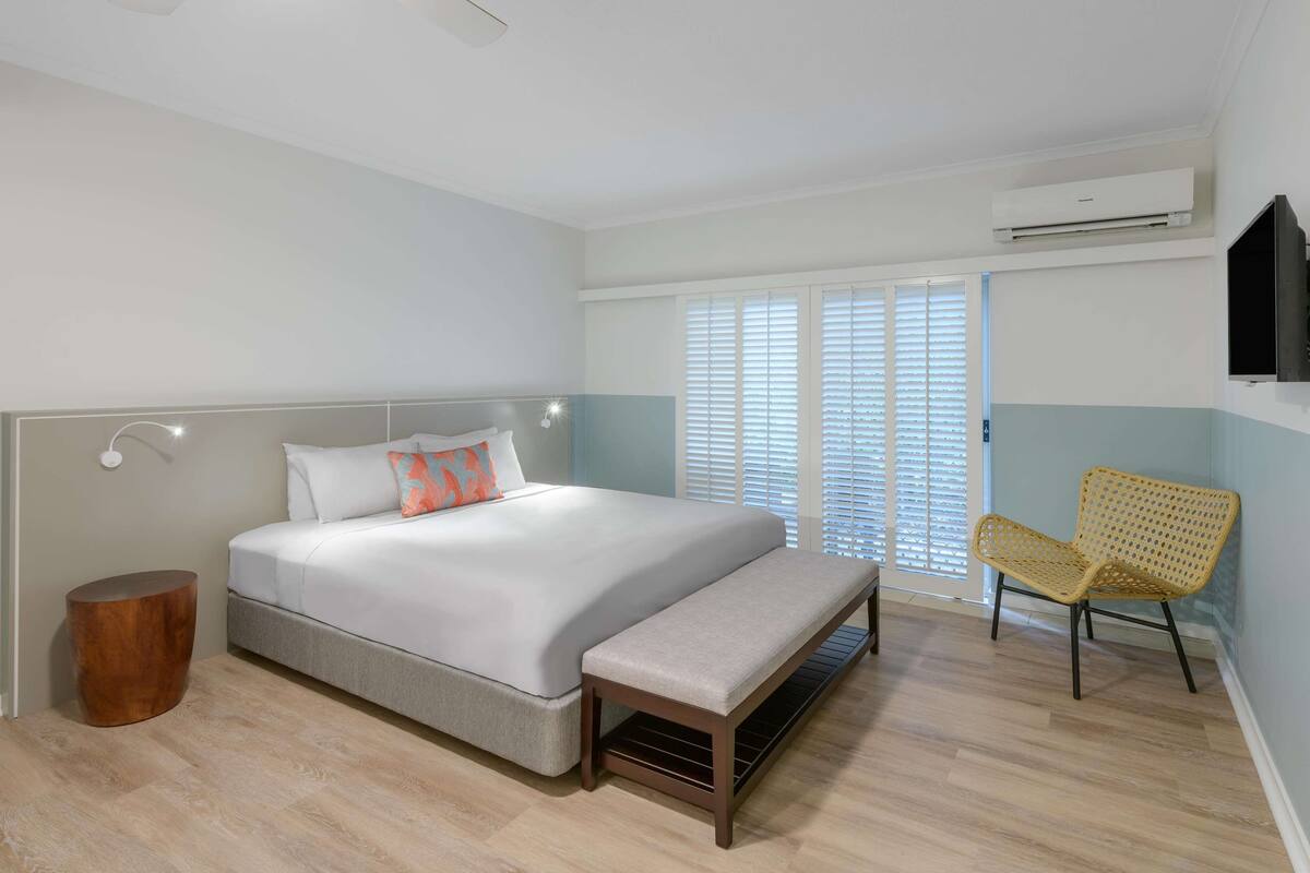 Un dormitorio moderno con una cama grande, un banco, una mesita redonda de madera, una silla de mimbre y un televisor montado en la pared.