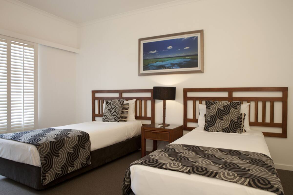 Habitación de hotel con dos camas individuales, una mesita de noche con lámpara, un cuadro en la pared y una ventana con persianas.