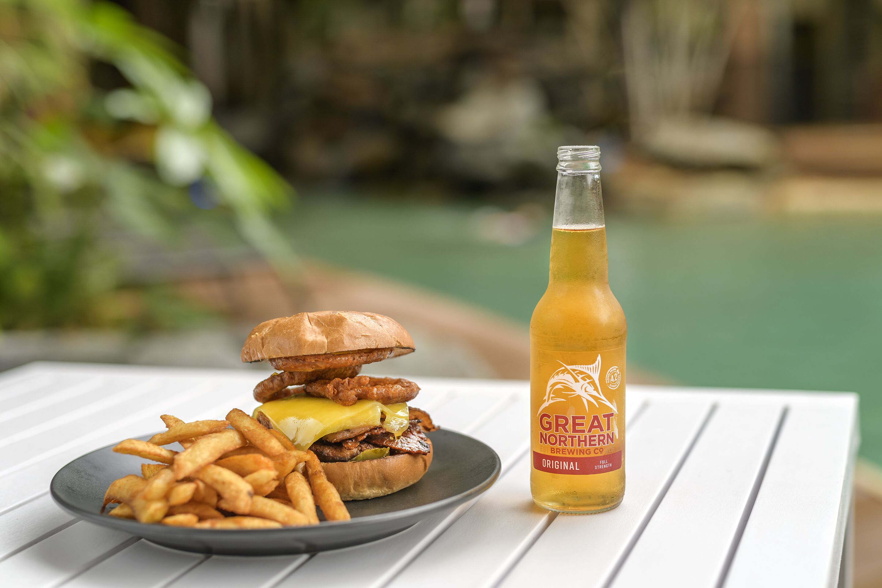 Een hamburger en frieten op een bord, naast een fles bier op een witte tafel.