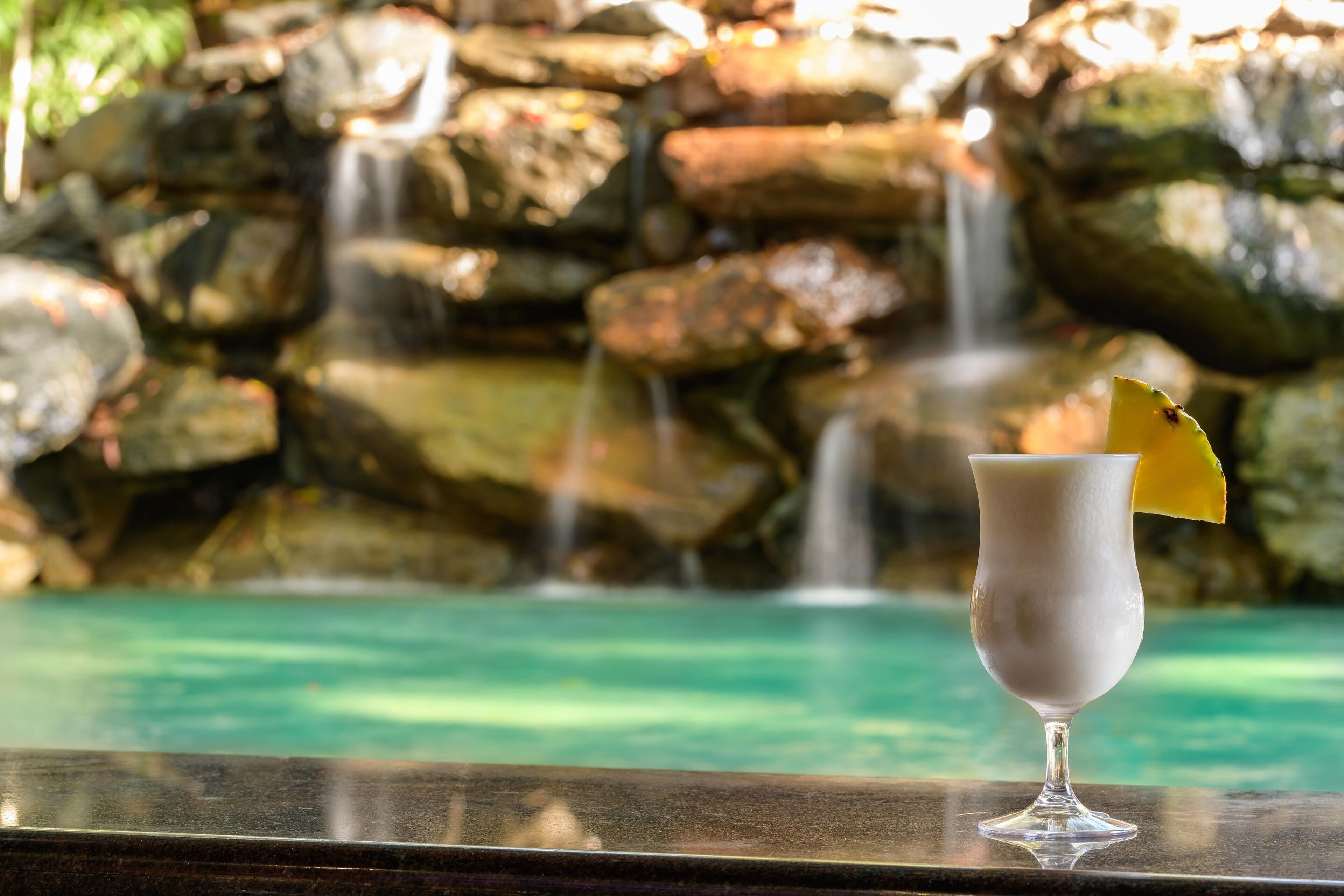 A glass of a white beverage with a yellow garnish on a bar counter, with a waterfall and rocky background.