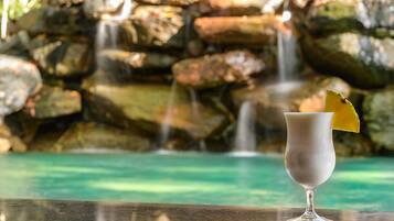 A glass of a white beverage with a yellow garnish on a bar counter, with a waterfall and rocky background.