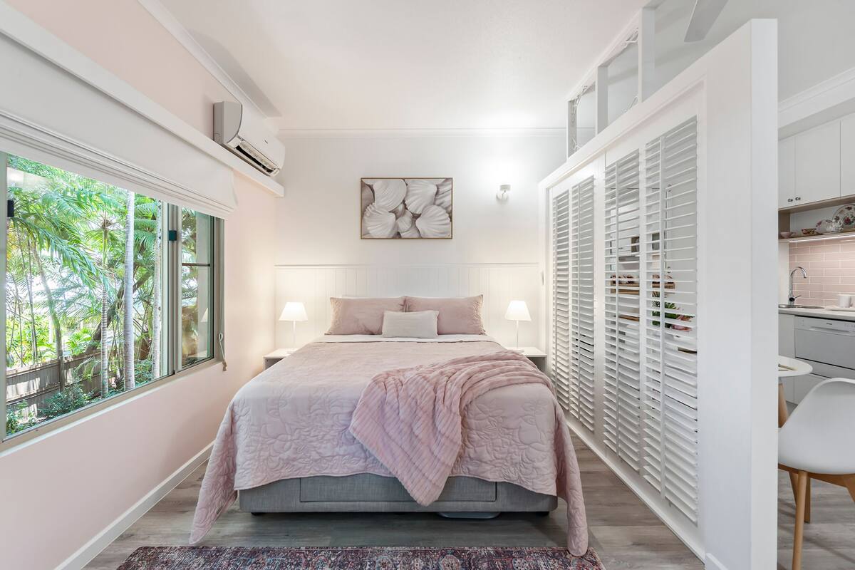 Un dormitorio con un ventanal grande, una cama con manta rosa, una mesita de noche con lámpara y un armario con contraventanas blancas.