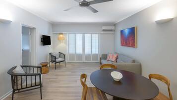 A modern living room with a dining area, a ceiling fan, a flat-screen TV, and a large window with blinds.