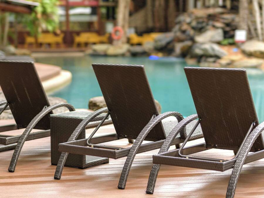 Three wicker lounge chairs with metal frames, positioned on a wooden deck by a pool with a rocky and plant-lined backdrop.