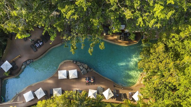 Luchtbeeld van een resort met een groot zwembad, omringd door loungegedeeltes en parasols, gelegen in een tropische omgeving met weelderige begroeiing.