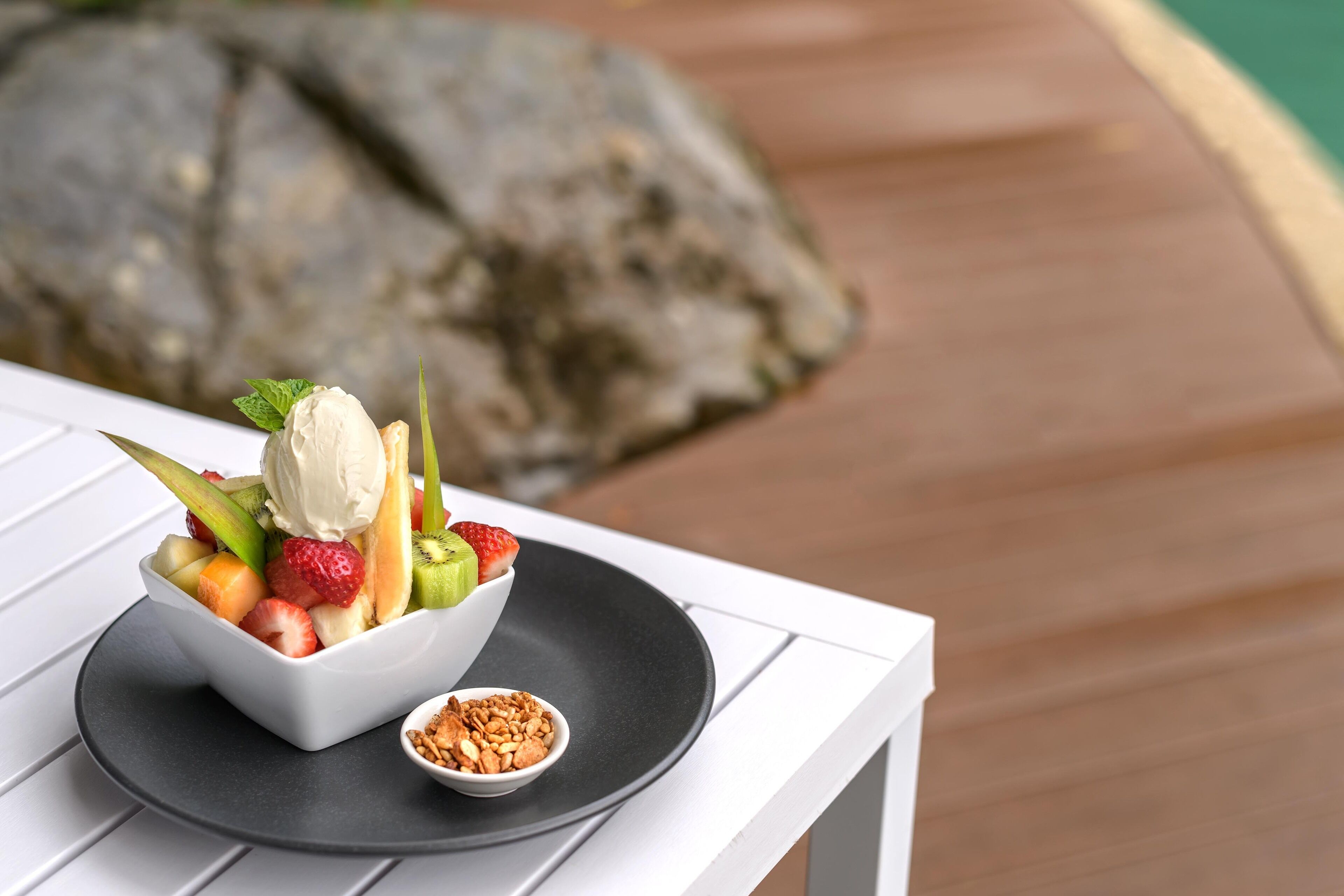 A dessert bowl with ice cream, strawberries, kiwi, and melon on a white table with a black plate and a small bowl of nuts.