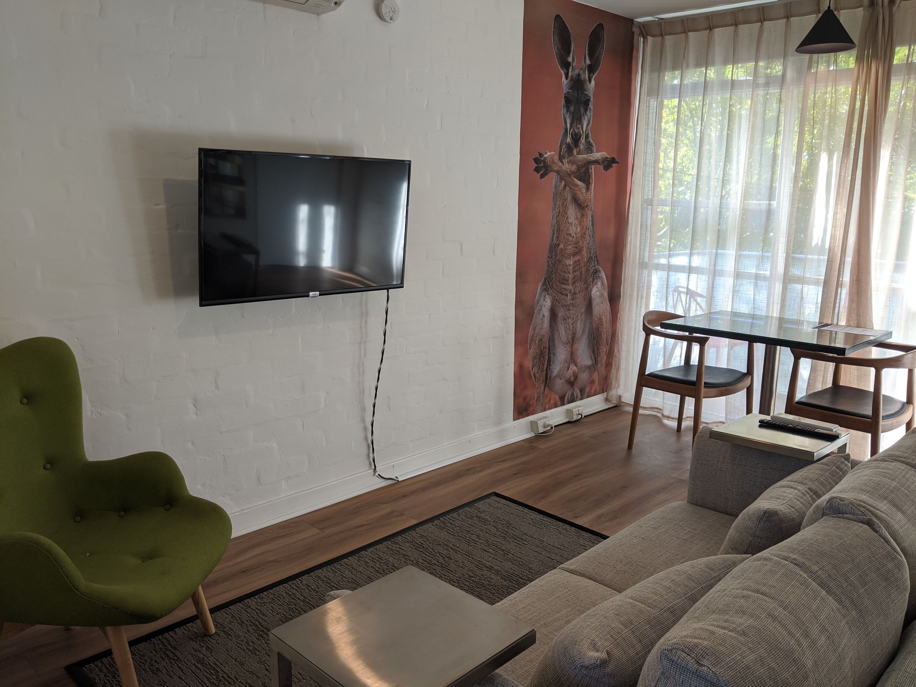 A living room with a flat-screen TV mounted on a white brick wall, a green armchair, a gray sofa, a glass dining table with wooden chairs, and a mural of a kangaroo.
