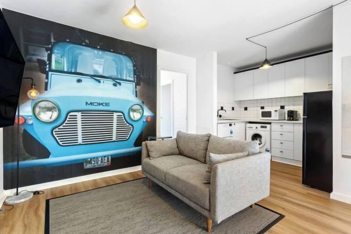 A modern living room with a sofa, a wall-mounted artwork of a blue car, and a kitchen in the background.