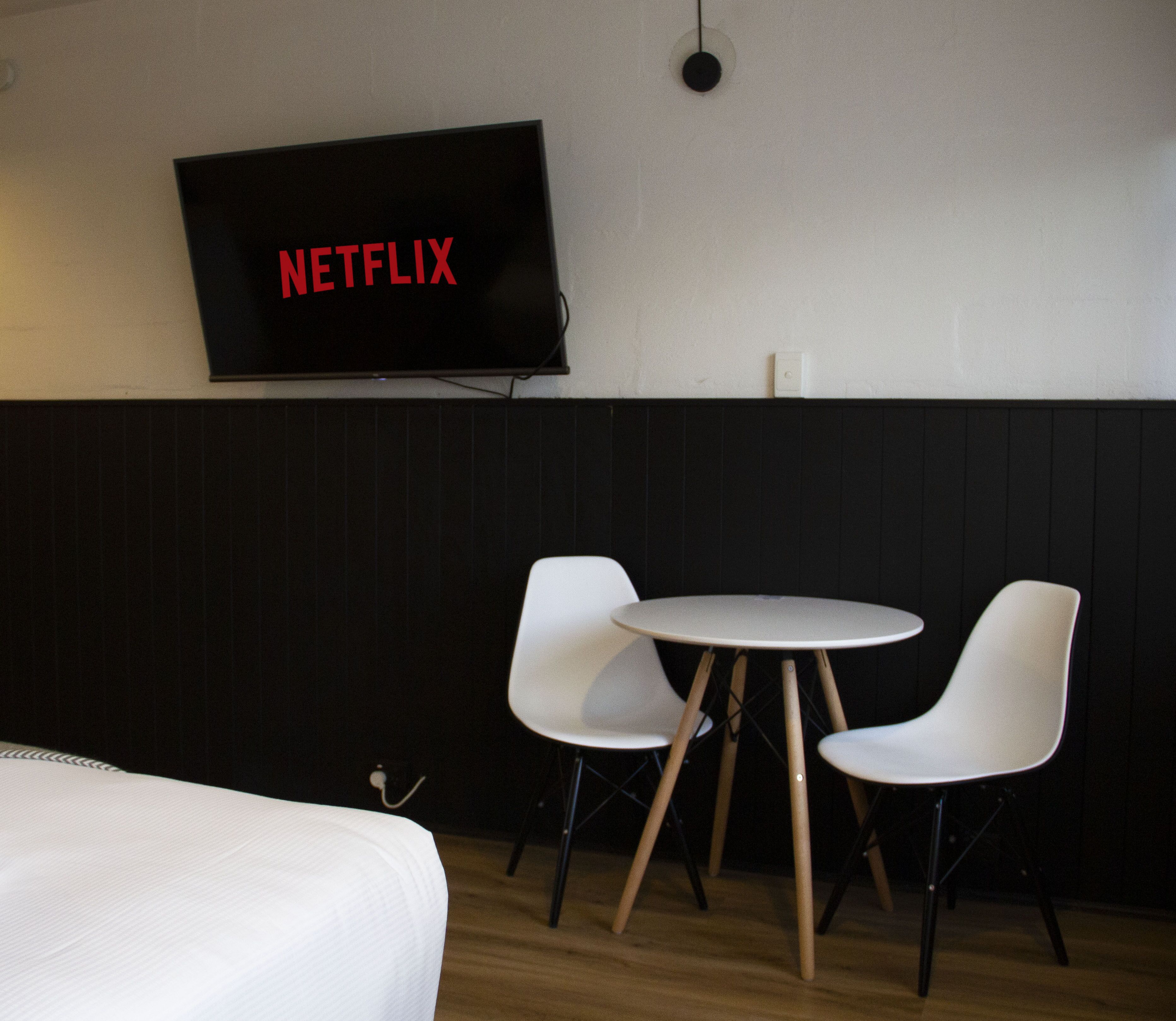 A room with a bed, a small round table with two chairs, a wall-mounted TV displaying "NETFLIX", and a wall switch.