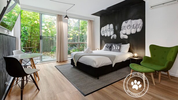 A modern bedroom with a large bed, a green armchair, a dining table with chairs, and a sliding glass door leading to a garden. - SoYa on Davis Apartment Hotel (South Yarra)