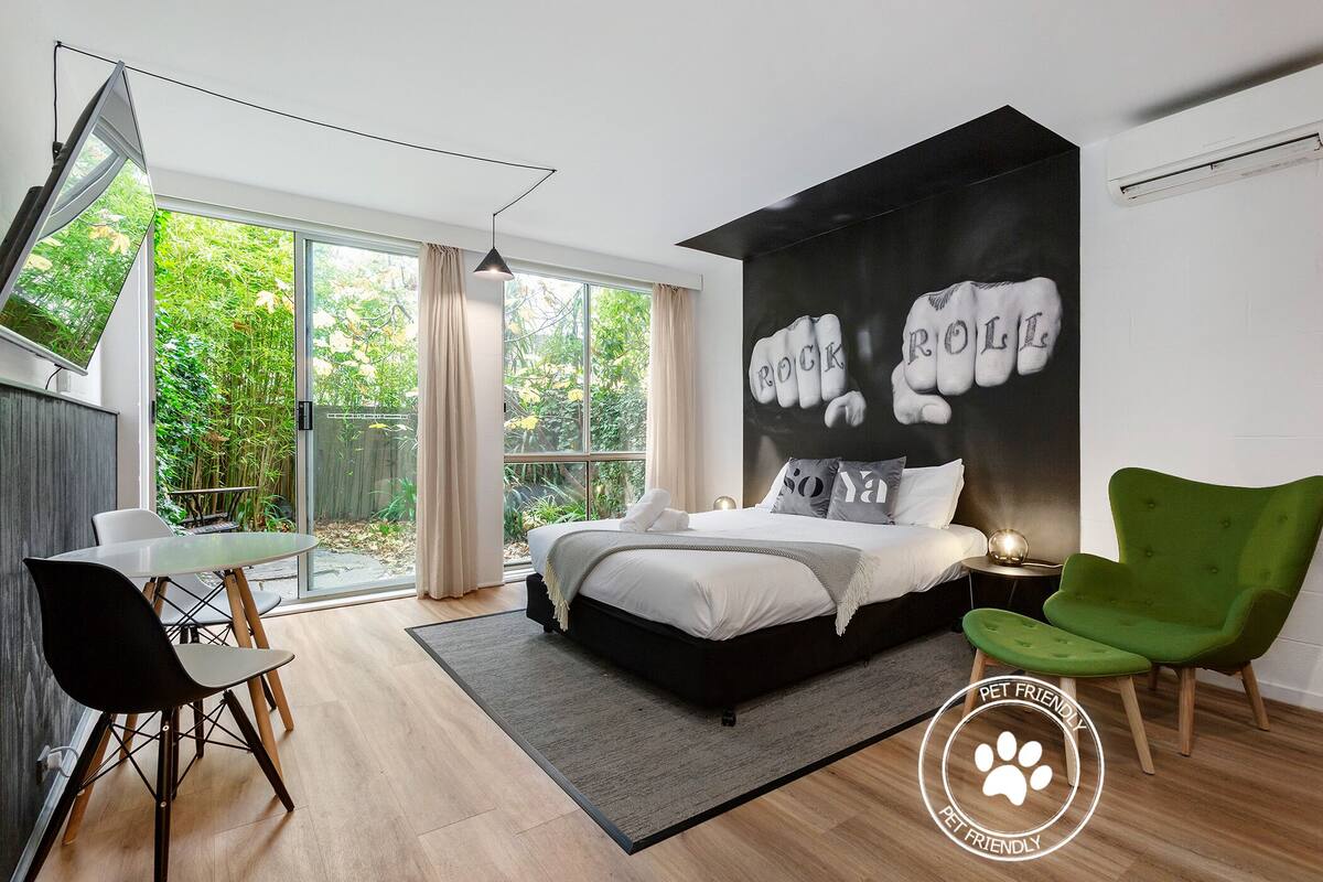 A modern bedroom with a large bed, a green armchair, a dining table with chairs, and a sliding glass door leading to a garden.
