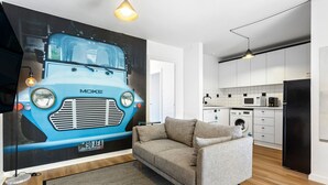 A modern living room with a sofa, a large wall mural of a blue bus, and a kitchen area with white cabinets and appliances. - SoYa on Davis Apartment Hotel (South Yarra)