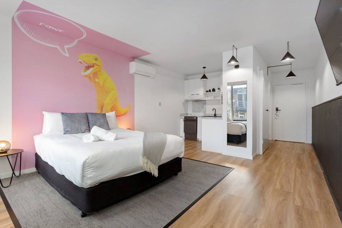 A modern bedroom with a bed, a pink wall with a dinosaur mural, a kitchen in the background, and pendant lights.
