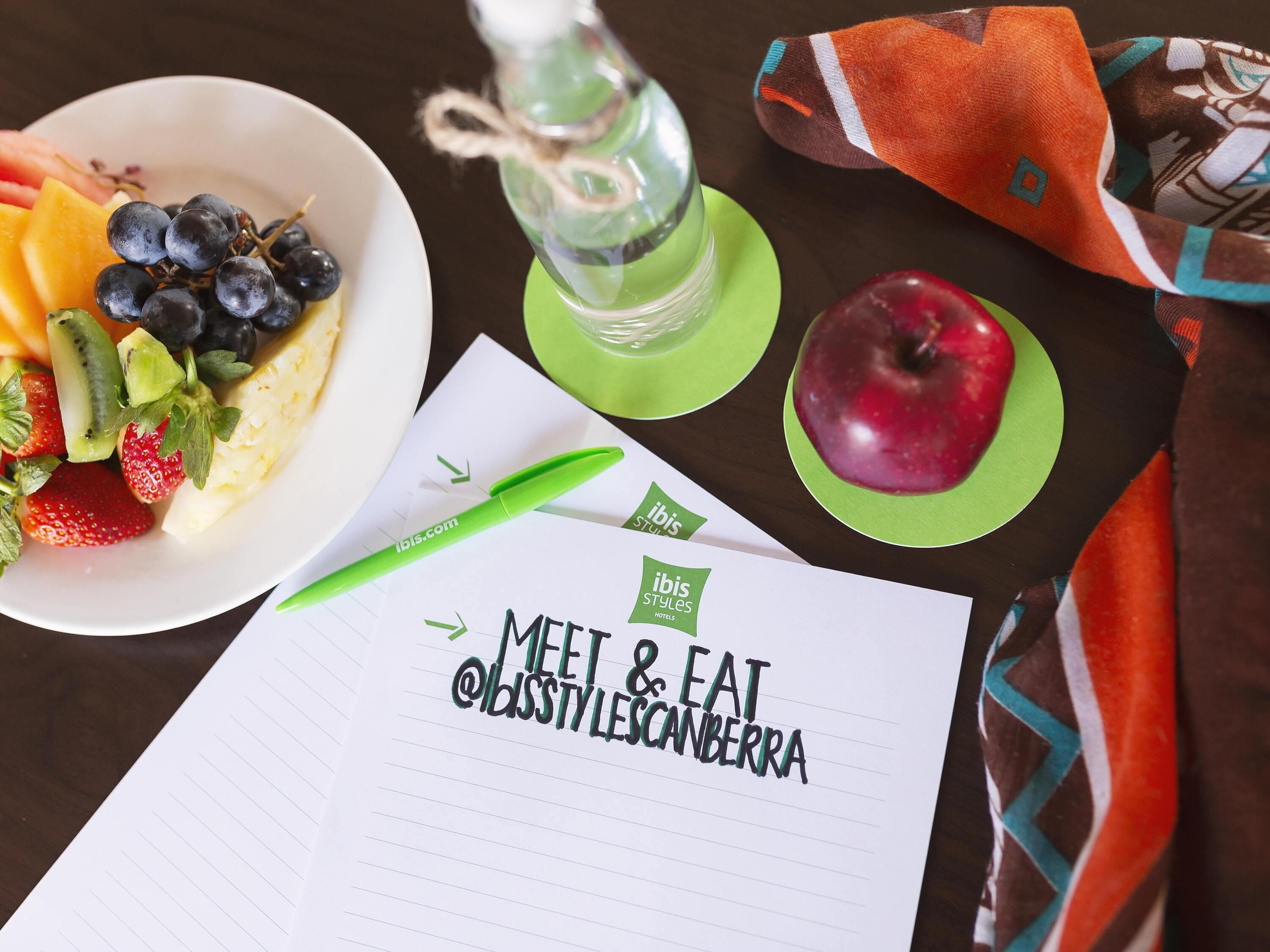 A table with a bowl of fruit, a green bottle, a red apple, and a notepad with hotel branding.