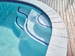 A clear swimming pool with blue mosaic tiles and a wooden deck surrounding it.
