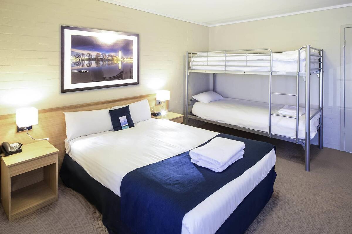 Una habitación de hotel con una cama grande, una litera, una mesita de noche con lámpara y un cuadro en la pared.