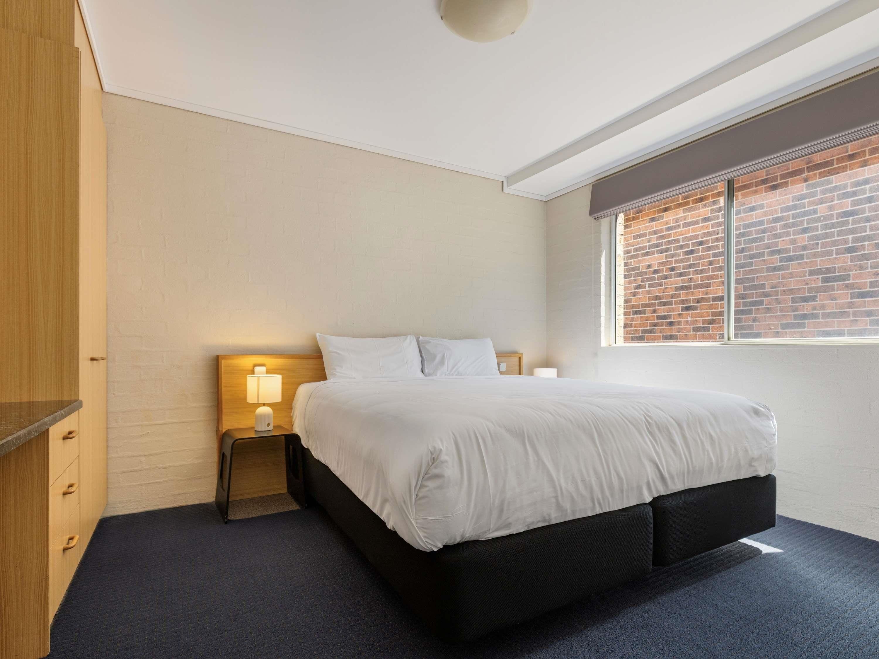 Un dormitorio con cama, mesitas de noche, una lámpara de techo y una ventana que da a un muro de ladrillo.