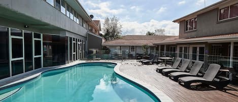 Una piscina con sillas de descanso, un porche cubierto y un edificio con amplios ventanales.
