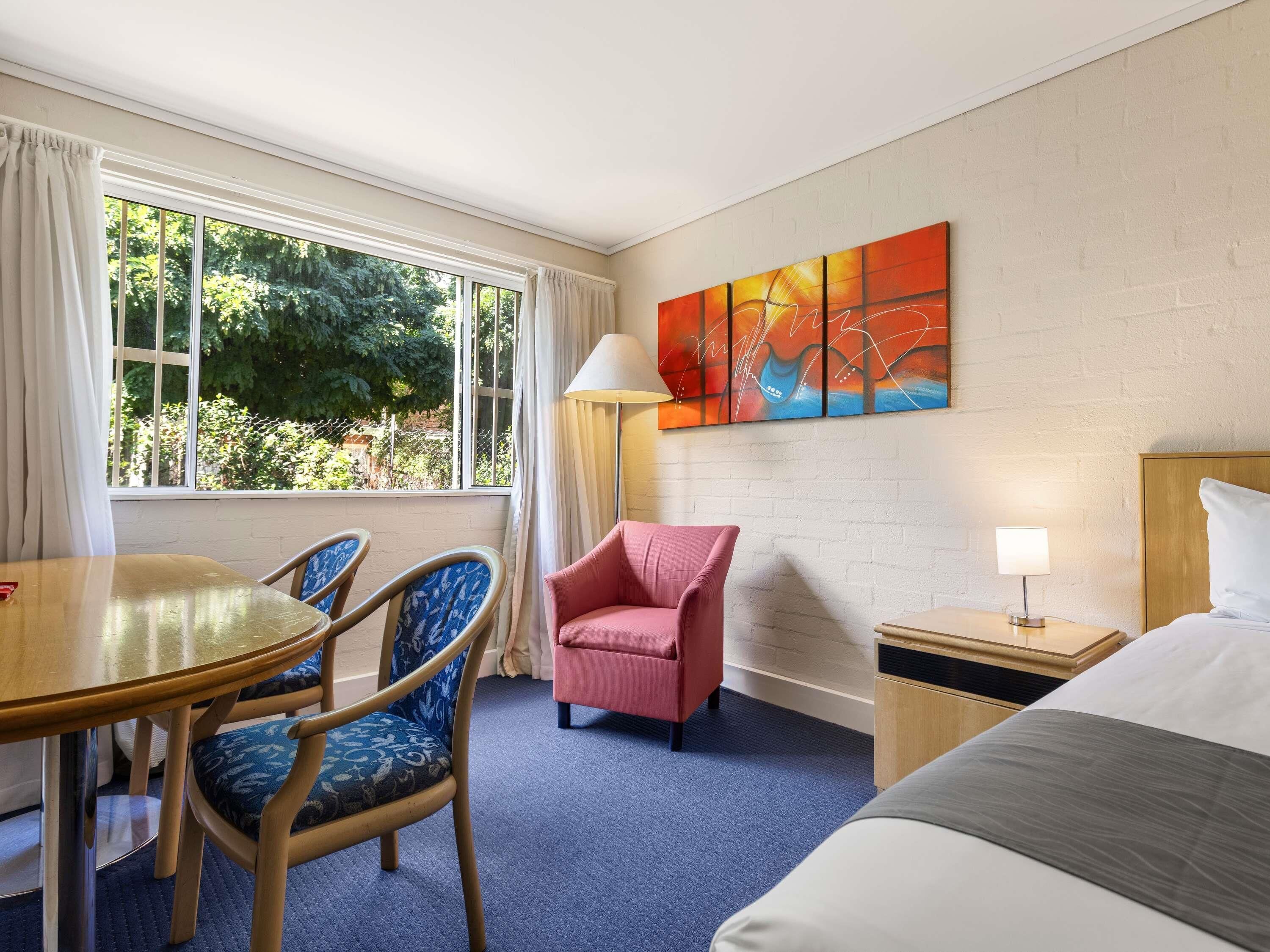 A hotel room with a bed, a table with chairs, a pink armchair, a lamp, and a painting on the wall.