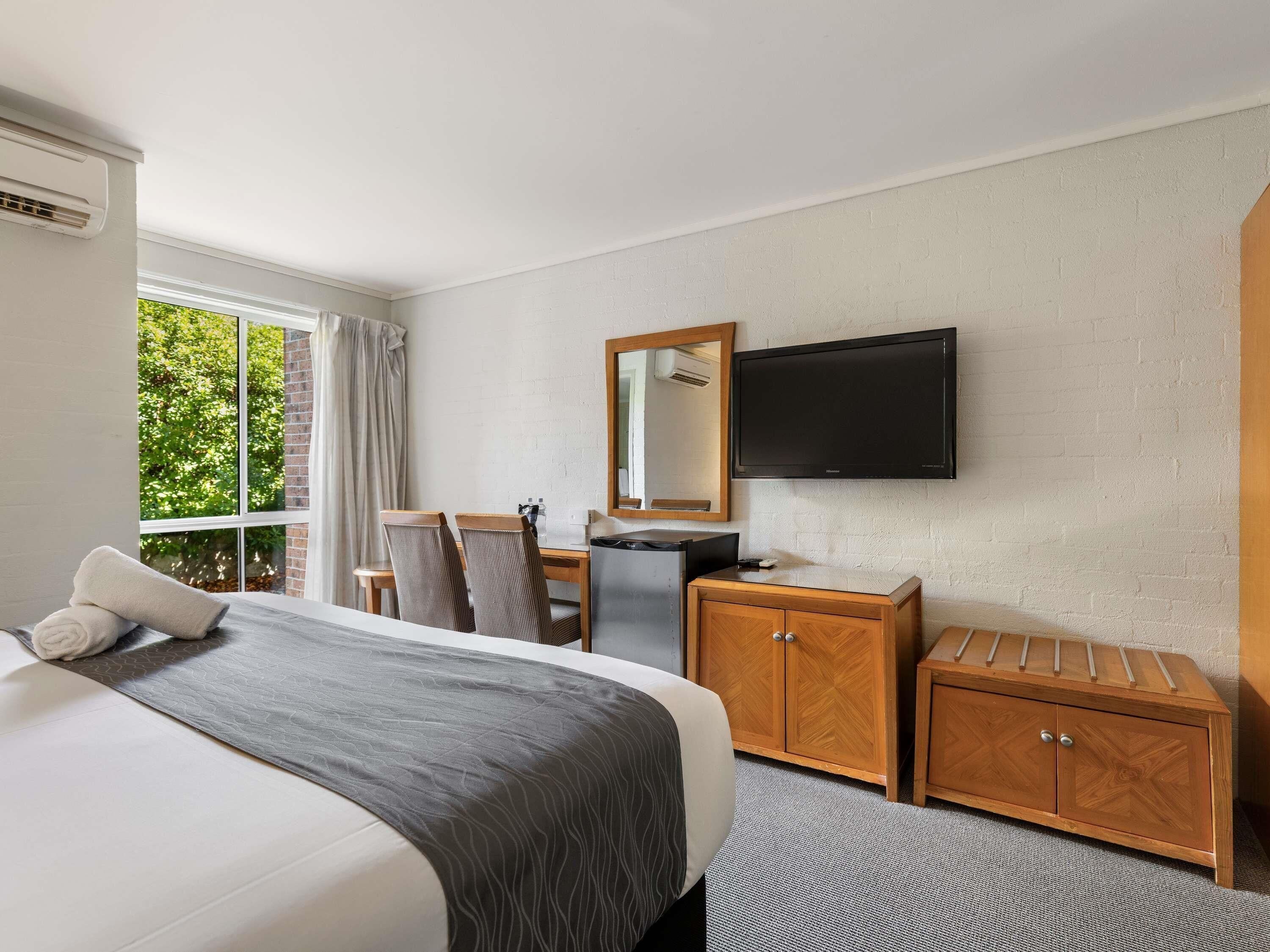 A hotel room with a bed, a desk, a mirror, a television, and a window with a view of greenery.