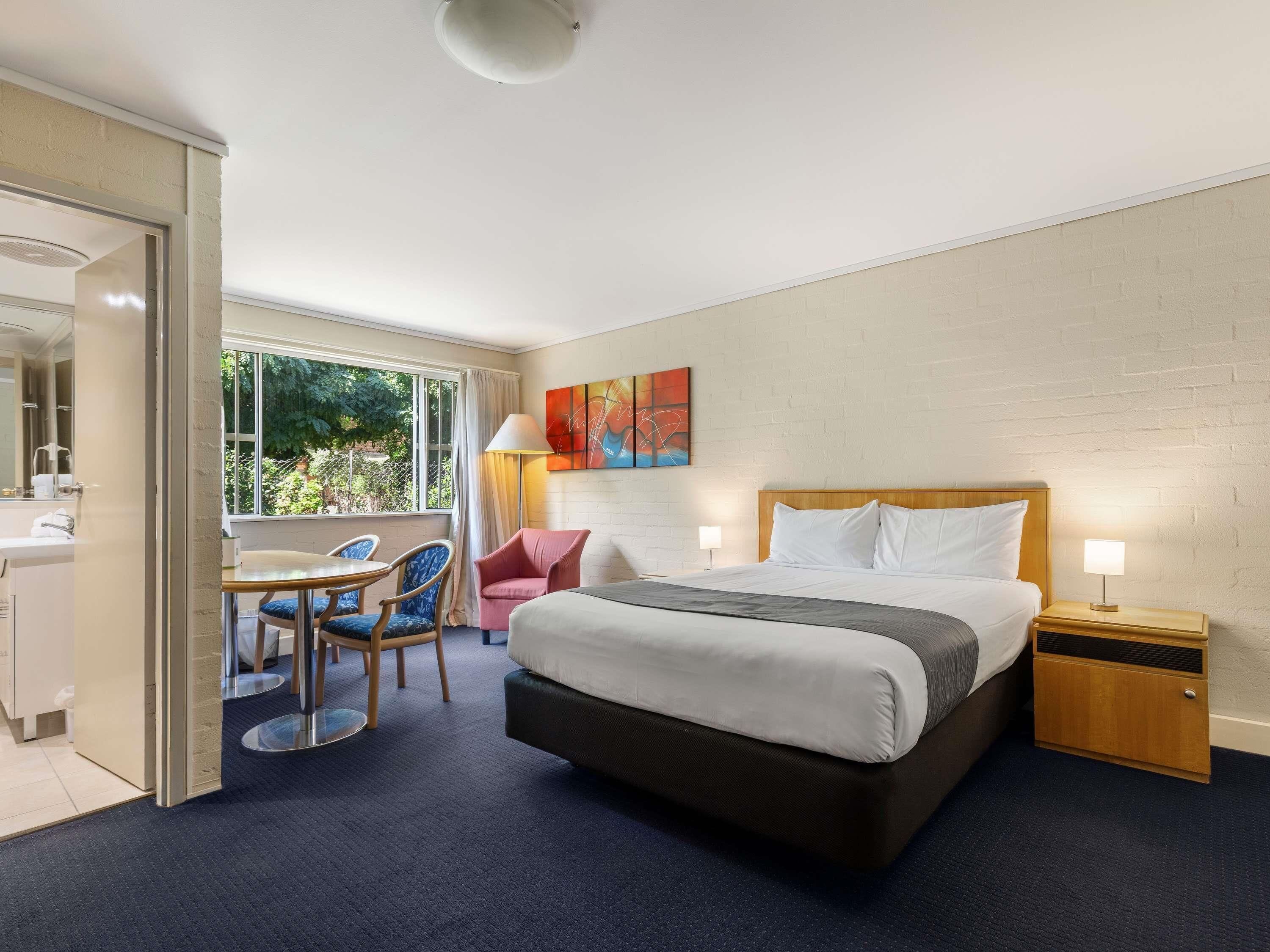 Habitación de hotel con cama, escritorio con silla, una mesita, lámpara y ventana con vista a zonas verdes.