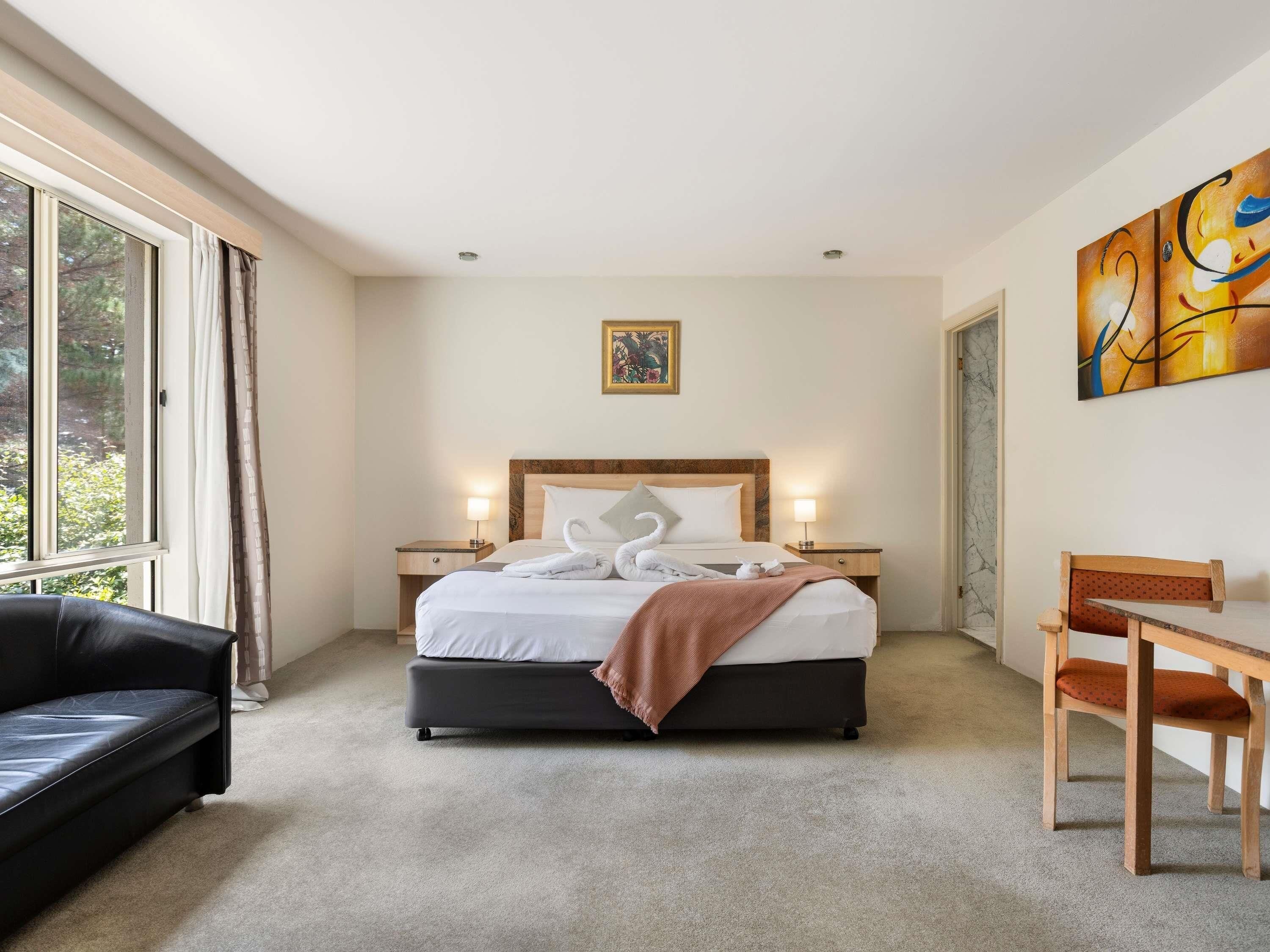 Habitación de hotel con una cama, dos mesitas de noche con lámparas, un sofá de cuero negro, una silla de madera y un amplio ventanal con cortinas.