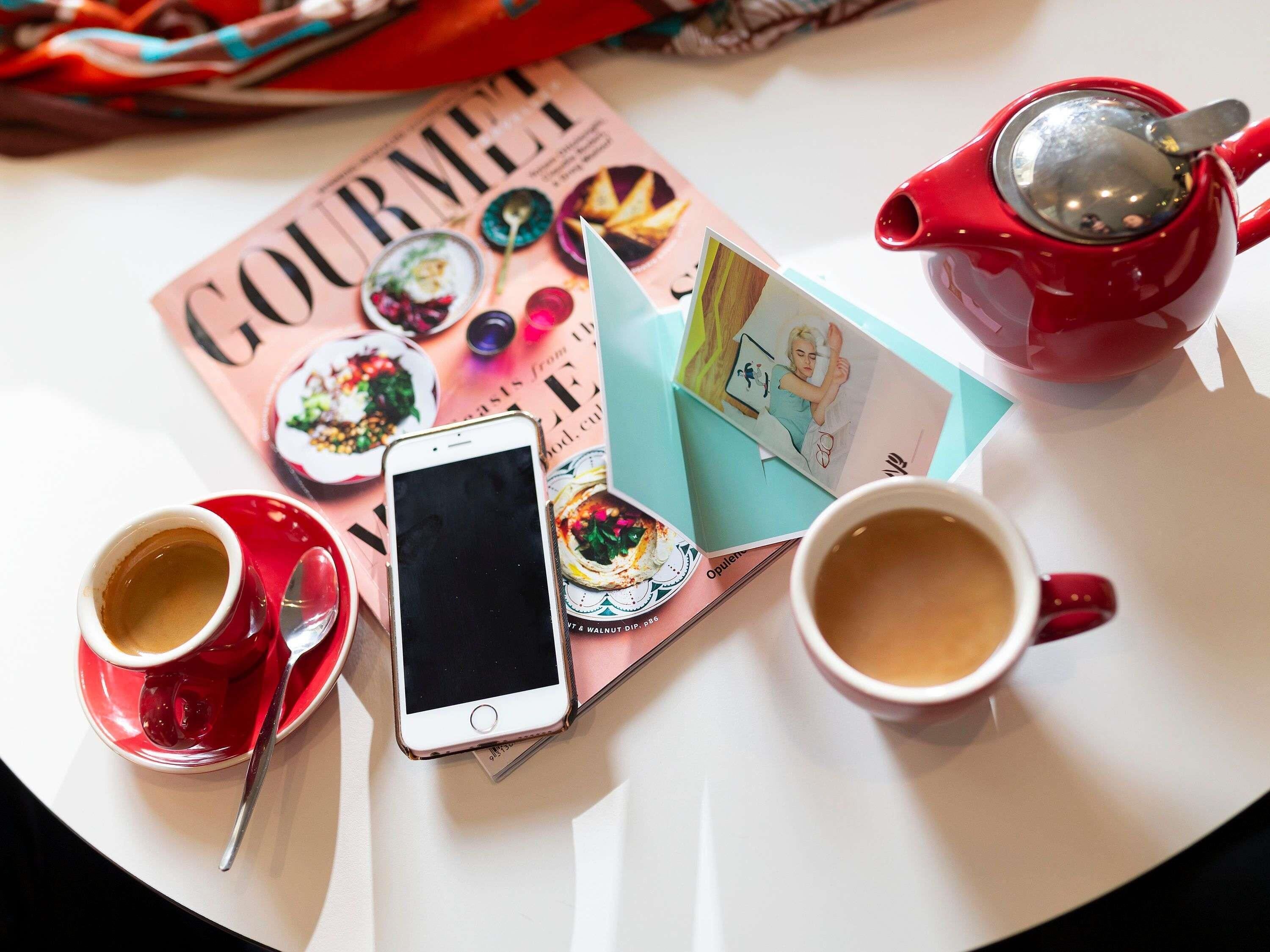 A table with a magazine, a smartphone, two cups of coffee, and a red teapot.
