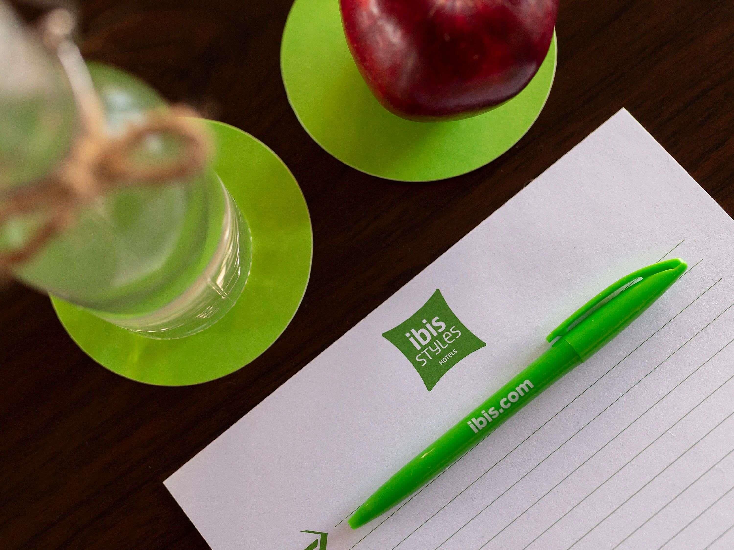 Un bolígrafo verde, una manzana roja y un vaso de agua sobre una mesa con un papel blanco que muestra el logo de 'ibis Styles Hotels'.