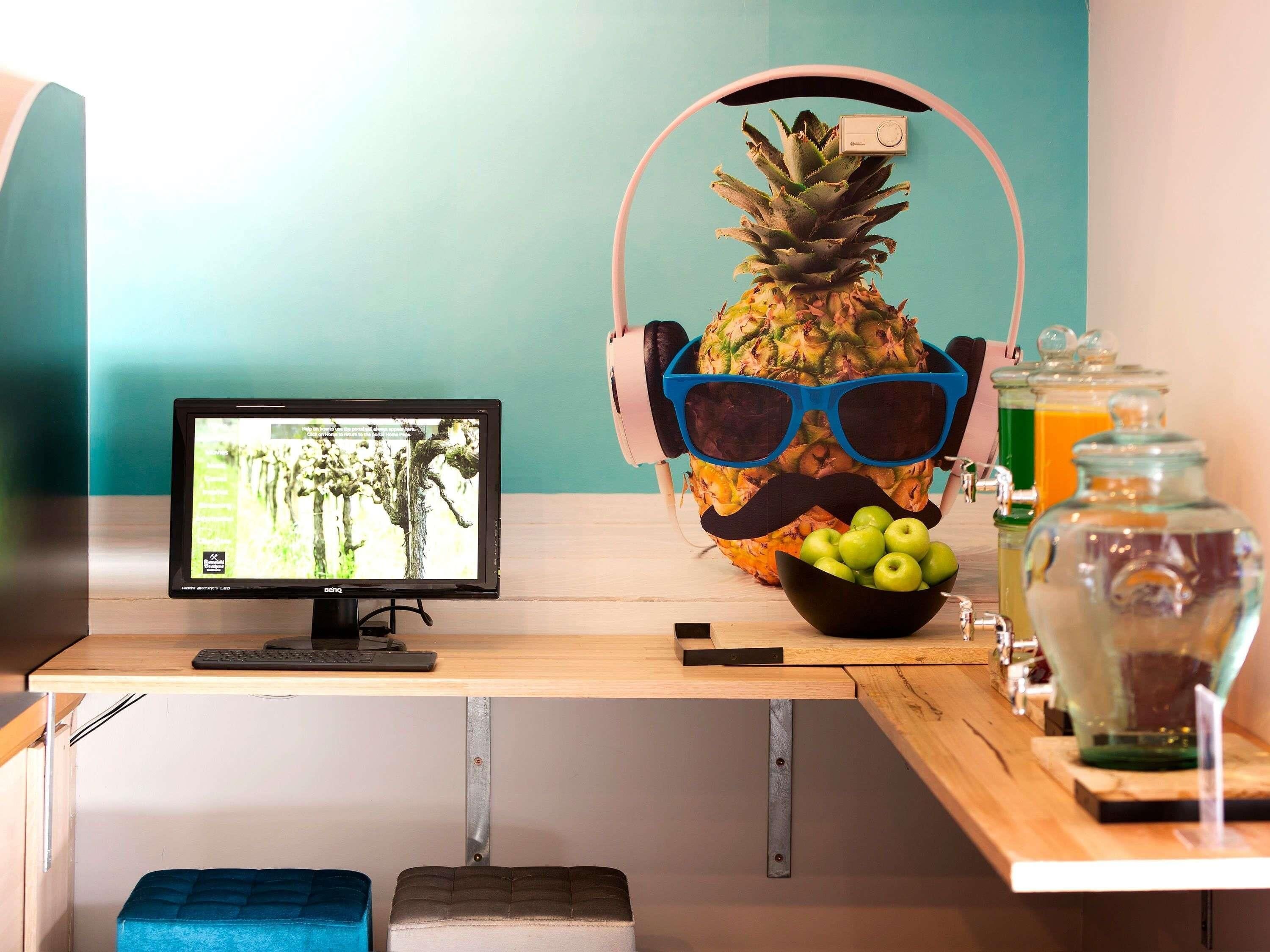 Un espacio de trabajo con un monitor de computadora, una piña con gafas de sol y bigote, un bol de manzanas y un dispensador de bebidas.