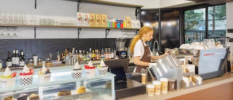 Interior de una cafetería con una vitrina de postres, una máquina de café y estantes con diversos artículos.