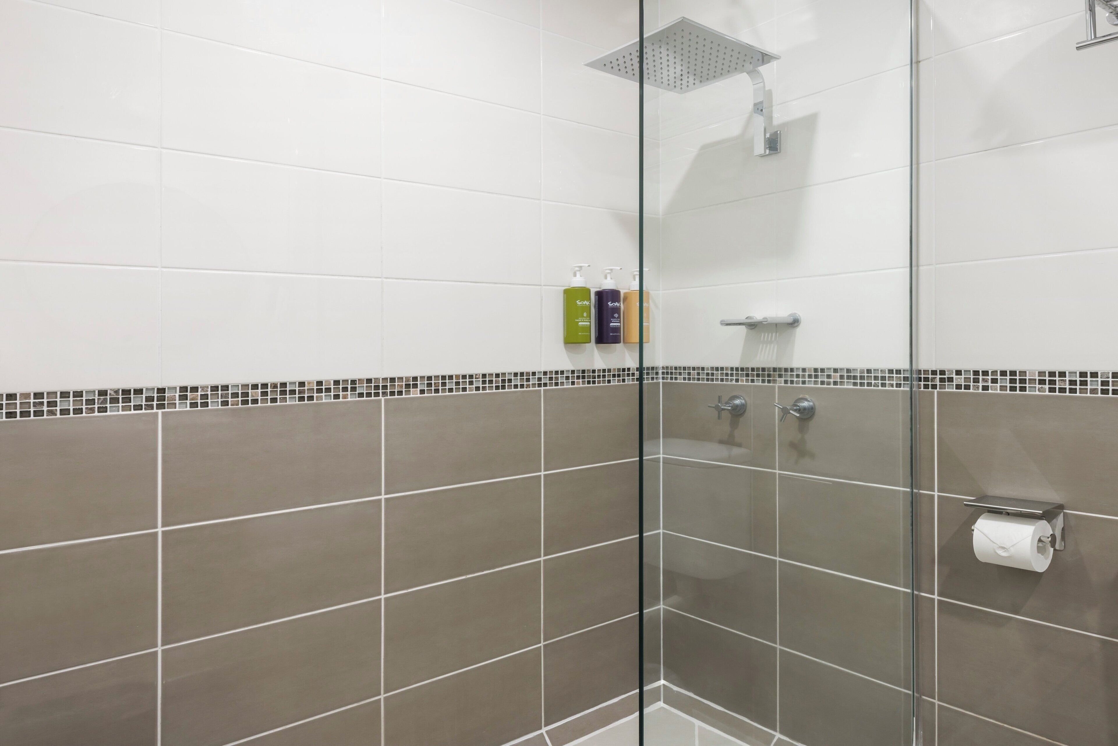 Un área de ducha con mampara de vidrio, cabezal de ducha manual y un porta papel higiénico.
