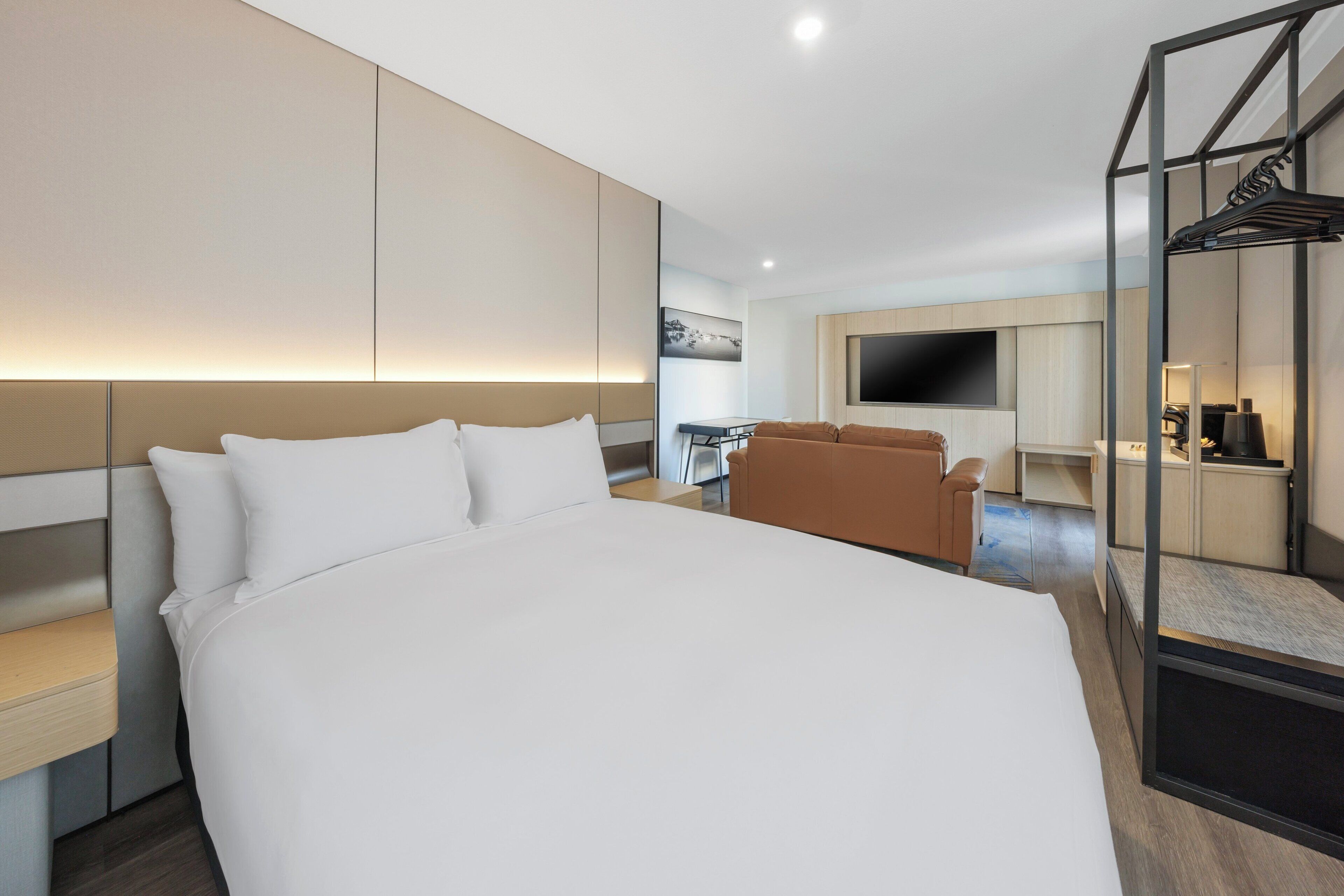 Quarto de hotel moderno com uma cama grande, área de estar com sofá, televisão de tela plana fixada na parede e espaço de jantar com mesa e cadeiras.