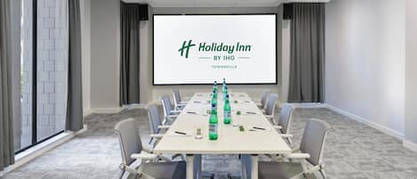 Una sala de conferencias con una pantalla grande mostrando el logotipo de Holiday Inn, una mesa larga con sillas y piso alfombrado.