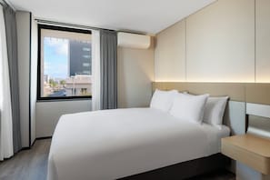 A hotel room with a large bed, a nightstand, a window with curtains, and a view of the cityscape.