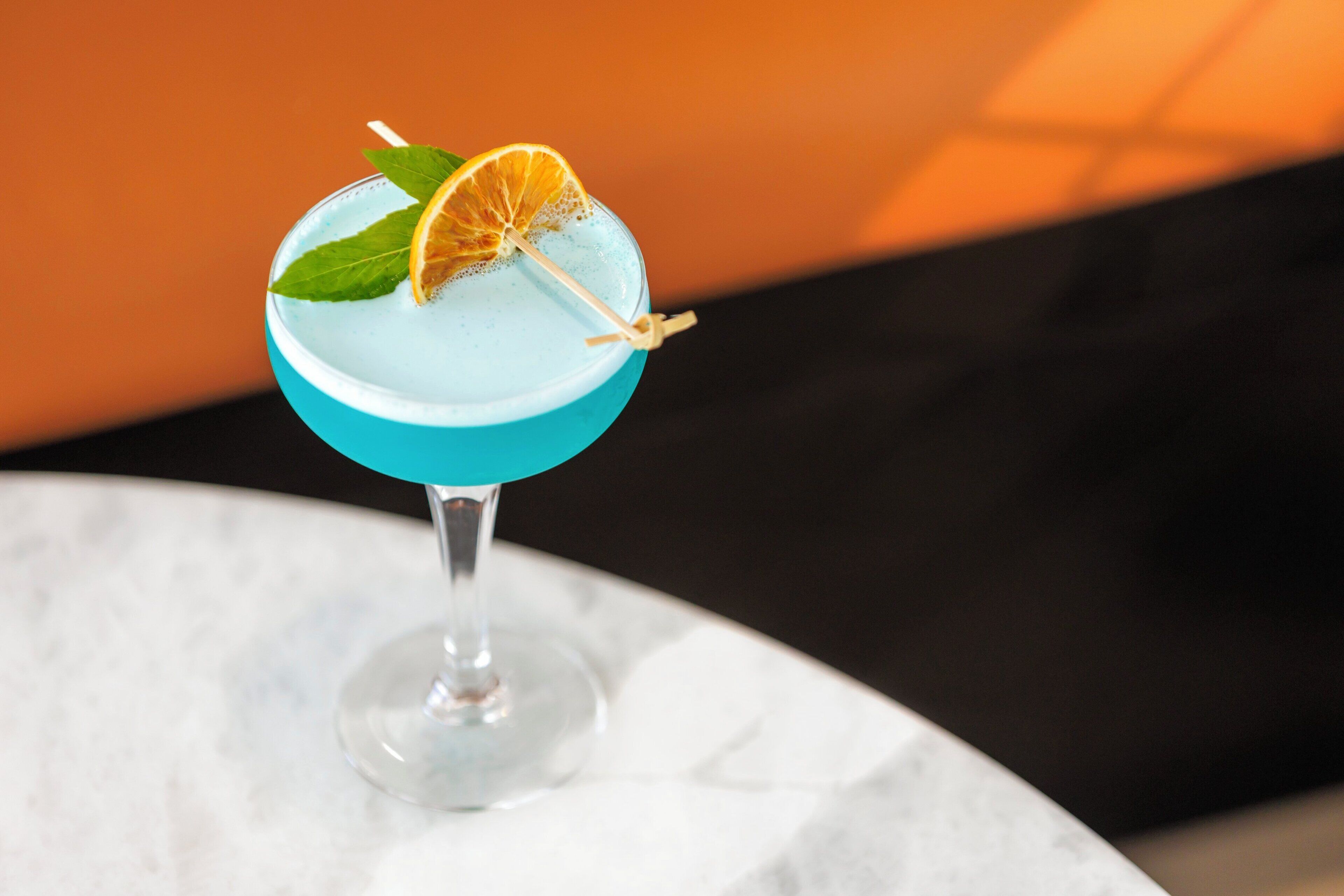 A cocktail in a blue glass with a slice of orange and a mint leaf garnish on a marble surface.