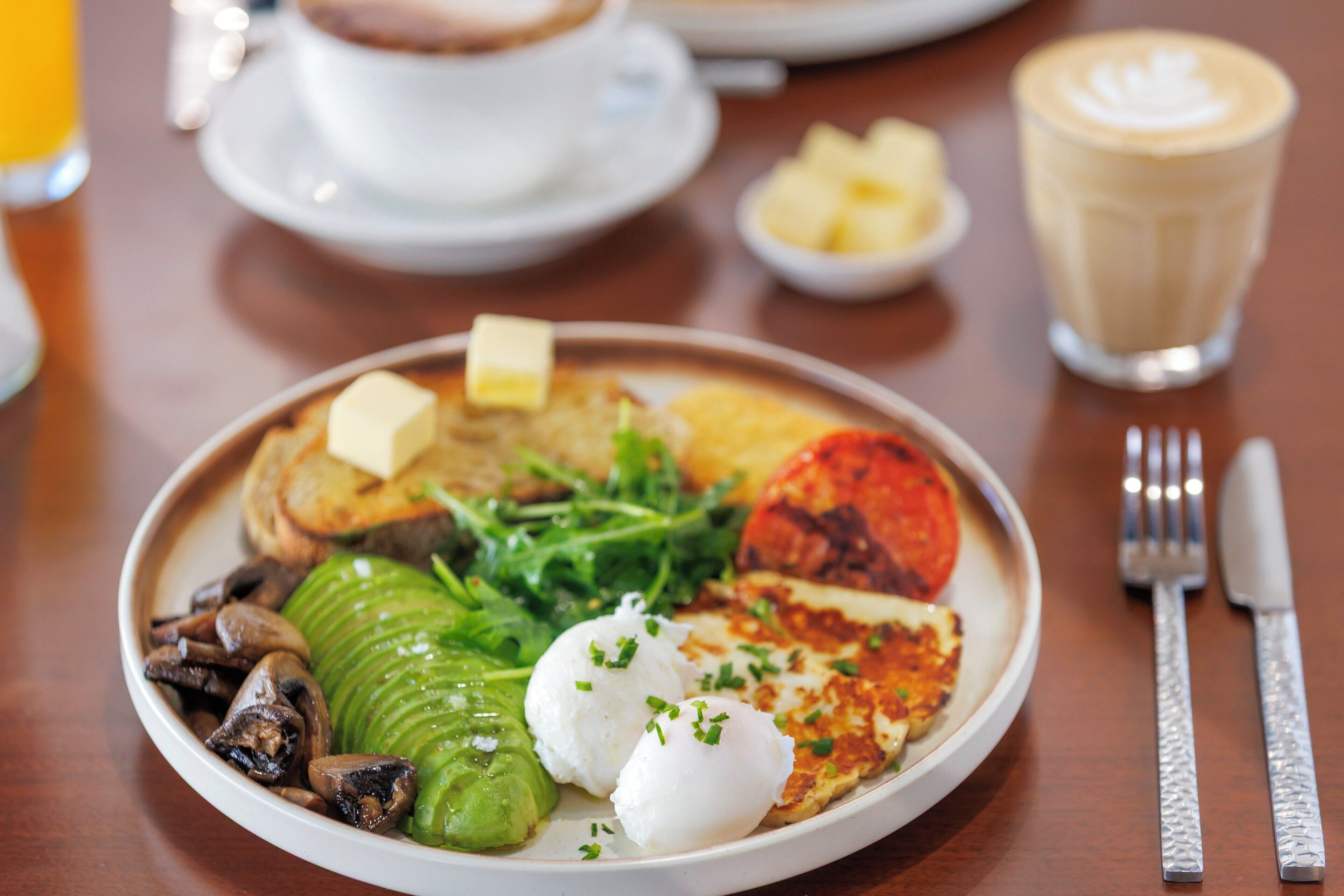 Un plato de desayuno con huevos, champiñones, verduras de hoja verde y tostadas, acompañado de una taza de café y un vaso de jugo de naranja.