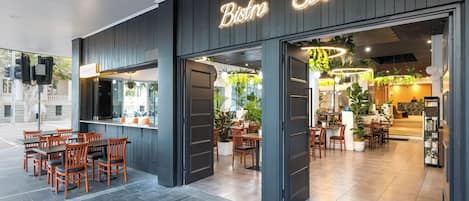 Un restaurante con letrero de neón, mesas al aire libre y un comedor interior con plantas.
