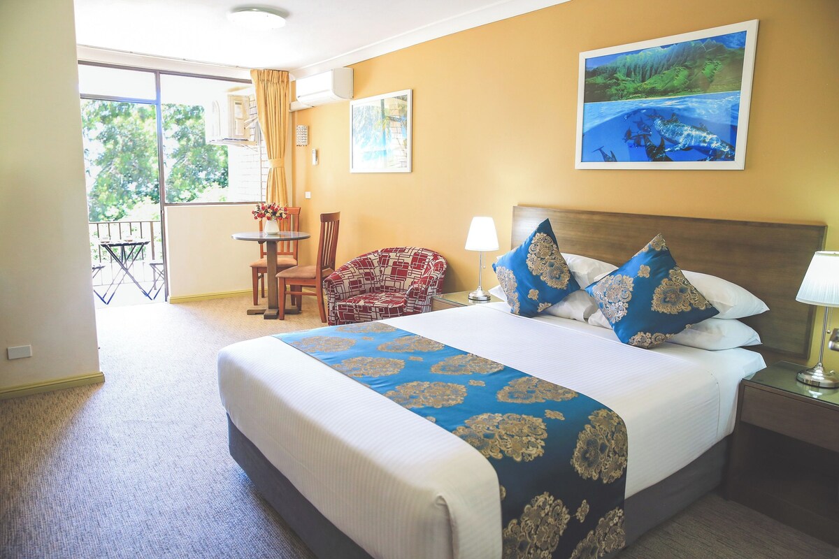 Een hotelkamer met een groot bed, een stoel, een tafel en een balkon met uitzicht op bomen.