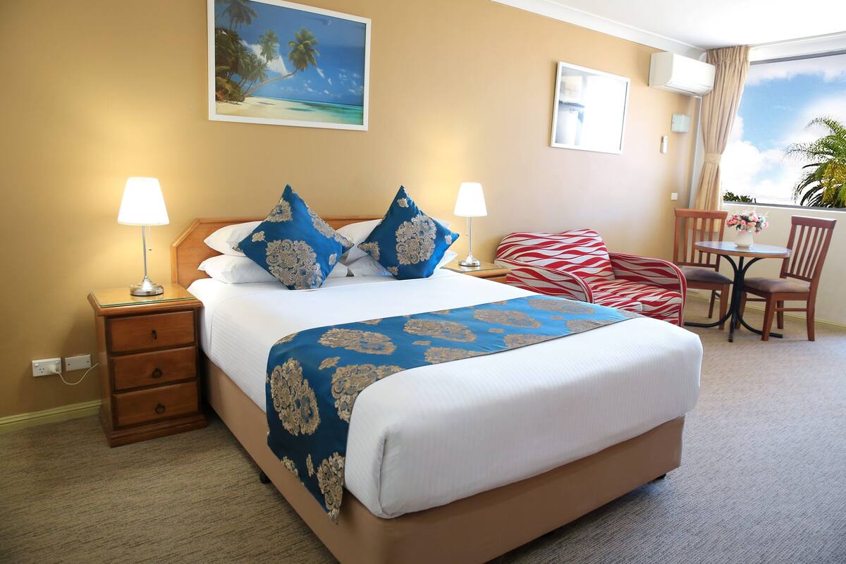 A hotel room with a large bed, two bedside tables with lamps, a small table with chairs, and a window with a view of palm trees.