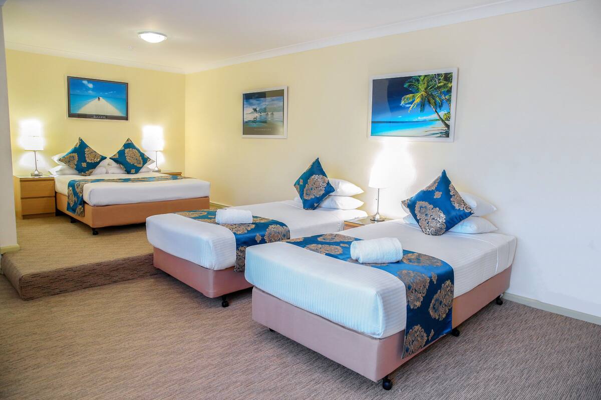 A hotel room with two beds, each with a blue and white bedspread, and matching pillows. There are two bedside tables with lamps, and the walls are decorated with framed pictures of tropical scenes.